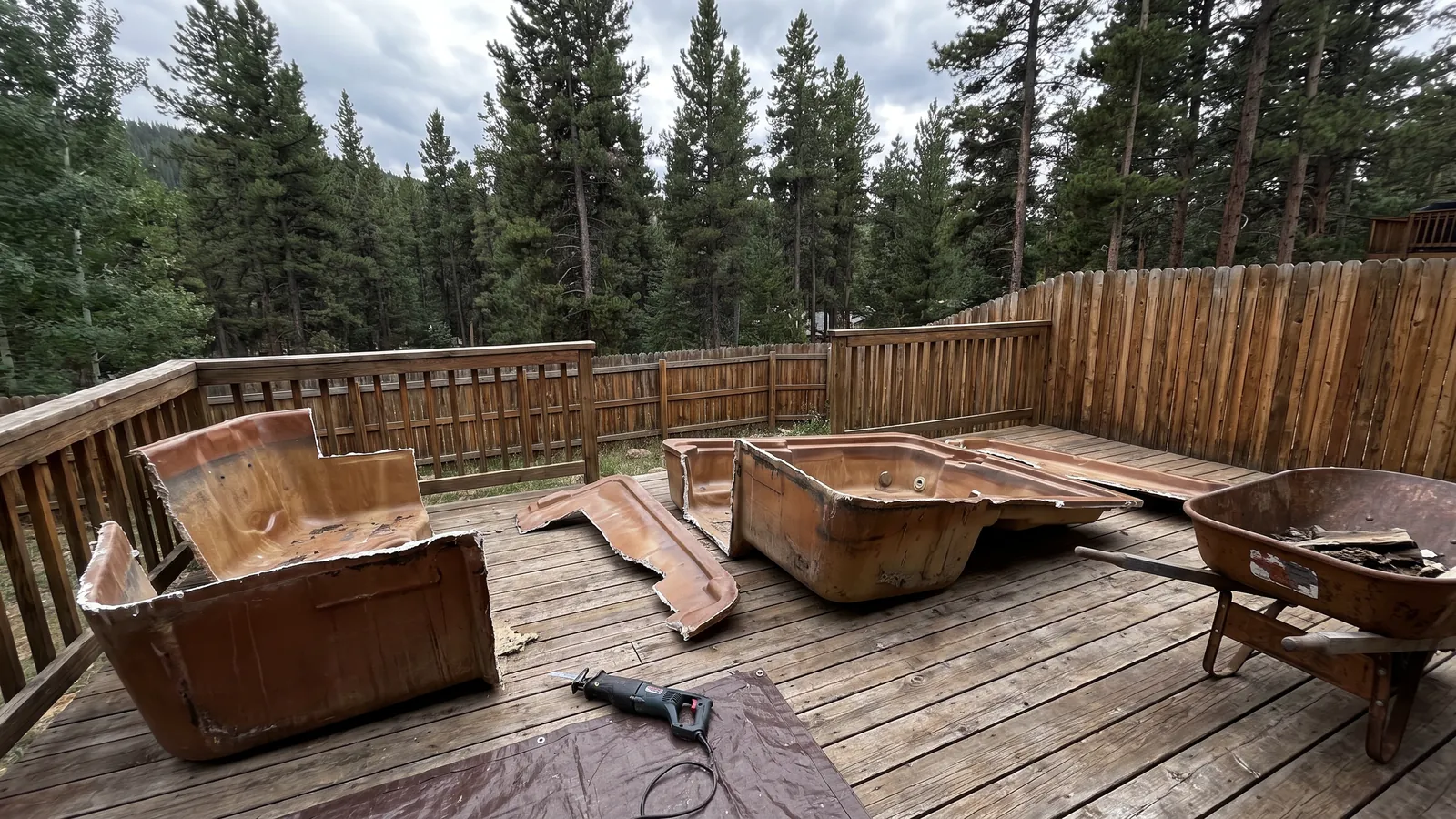 An old acrylic hot tub cut into sections on a backyard deck, a reciprocating saw and a wheelbarrow beside the pieces, pine trees behind the fence